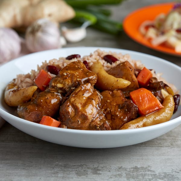 A white bowl of Jamaican brown stew chicken with rice, beans, carrots, and potatoes.