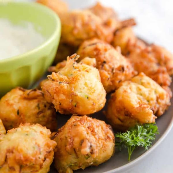 Golden-brown Jamaican fritters, possibly ackee or saltfish, with a side of white dipping sauce and parsley.