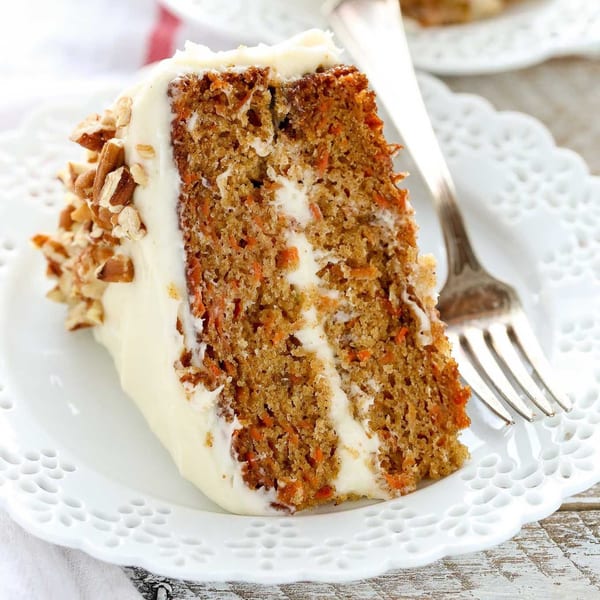 A slice of carrot cake with white frosting and chopped pecans on a white doily plate.